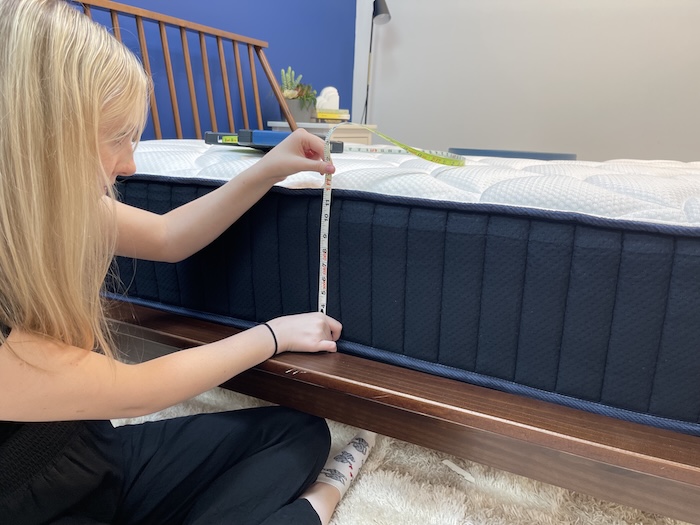 A woman sitting next to the DreamCloud mattress and using a tape measure to measure the bed's height.