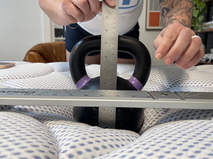 A close-up of a kettlebell resting on the DreamCloud Premier Hybrid mattress. Someone is measuring the sinkage of the bed.