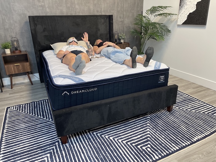 Two women lying next to each other on the DreamCloud Premier Hybrid mattress. One of them wears an eye mask and is raising her hand to indicate that she can feel her partner's movement.