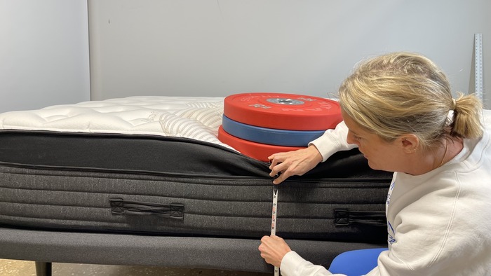 A woman kneeling next to the Nolah Evolution mattress while it has a stack of weights on it. She's measuring the bed's sinkage.