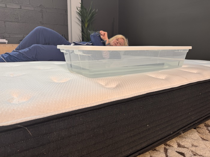 A woman lying on the Plank Firm mattress next to a plastic bin full of water. She's rolling around to test out the bed's motion isolation.