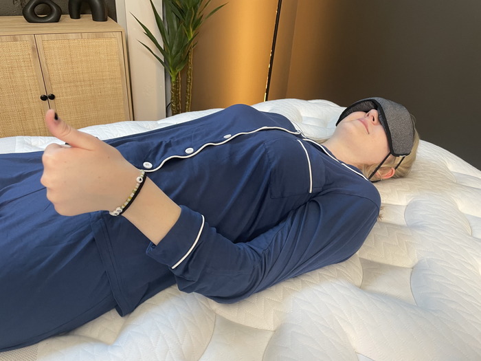 A woman lying on the Leesa Sapira Chill Hybrid. She's wearing an eye mask and giving a thumbs-up to indicate that she can feel her partner moving around.