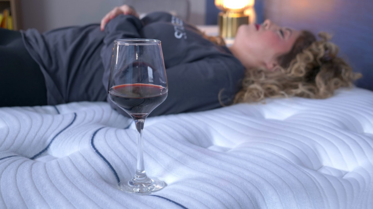 A woman lying down on a mattress next to a glass of red wine to test the bed's motion isolation.
